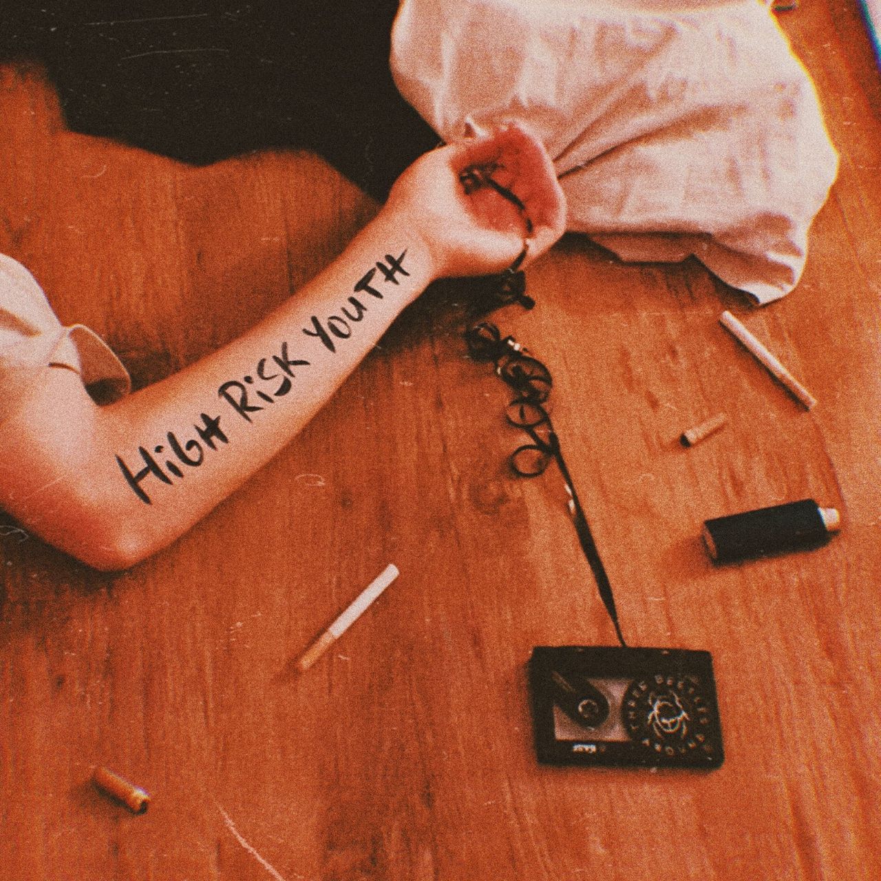 An arm of a person lying on the ground holding the tangled tape of a cassette with the TBA logo on it, surrounded by cigarrette buds.